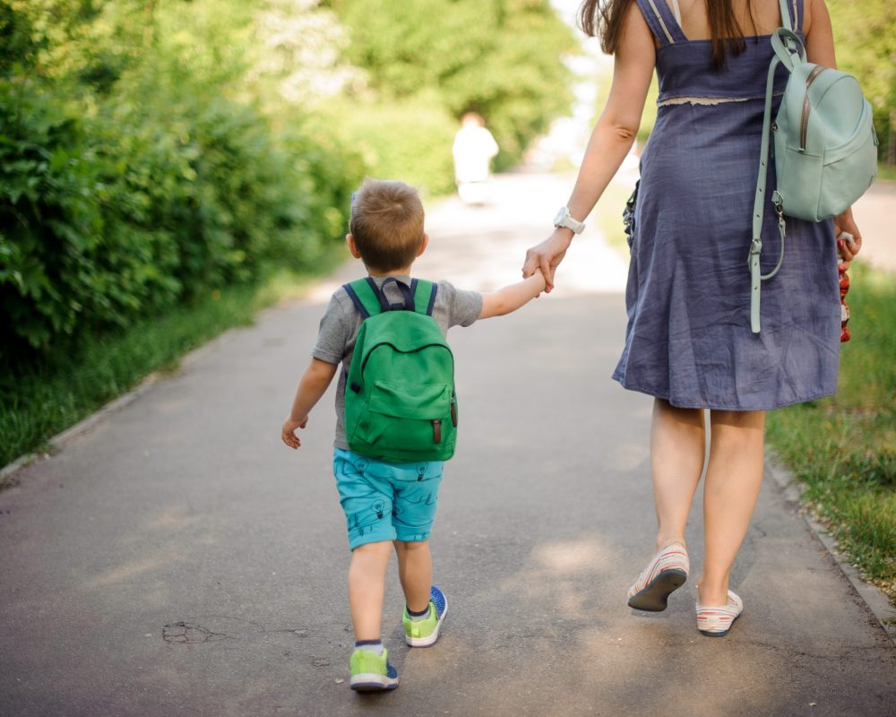 Back view of mother walking down the street with a little son with a backpack on sunny summer day